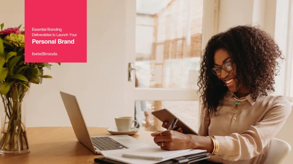 A person sitting at a table with a laptop, tablet, notebook, and coffee cup, working or studying. A vase with flowers is on the table, and natural light enters through a glass-paneled door.